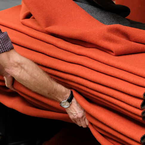 Ein älterer Mann legt mehrere Merinodecken »FireSideMoon« der Marke COASTLAND übereinander. Die Decken sind in glutrotem und anthrazitfarbenem Wollstoff gehalten und wirken weich und edel. Im Hintergrund sind weitere Decken zu sehen, die sich elegant über eine Vorrichtung legen.