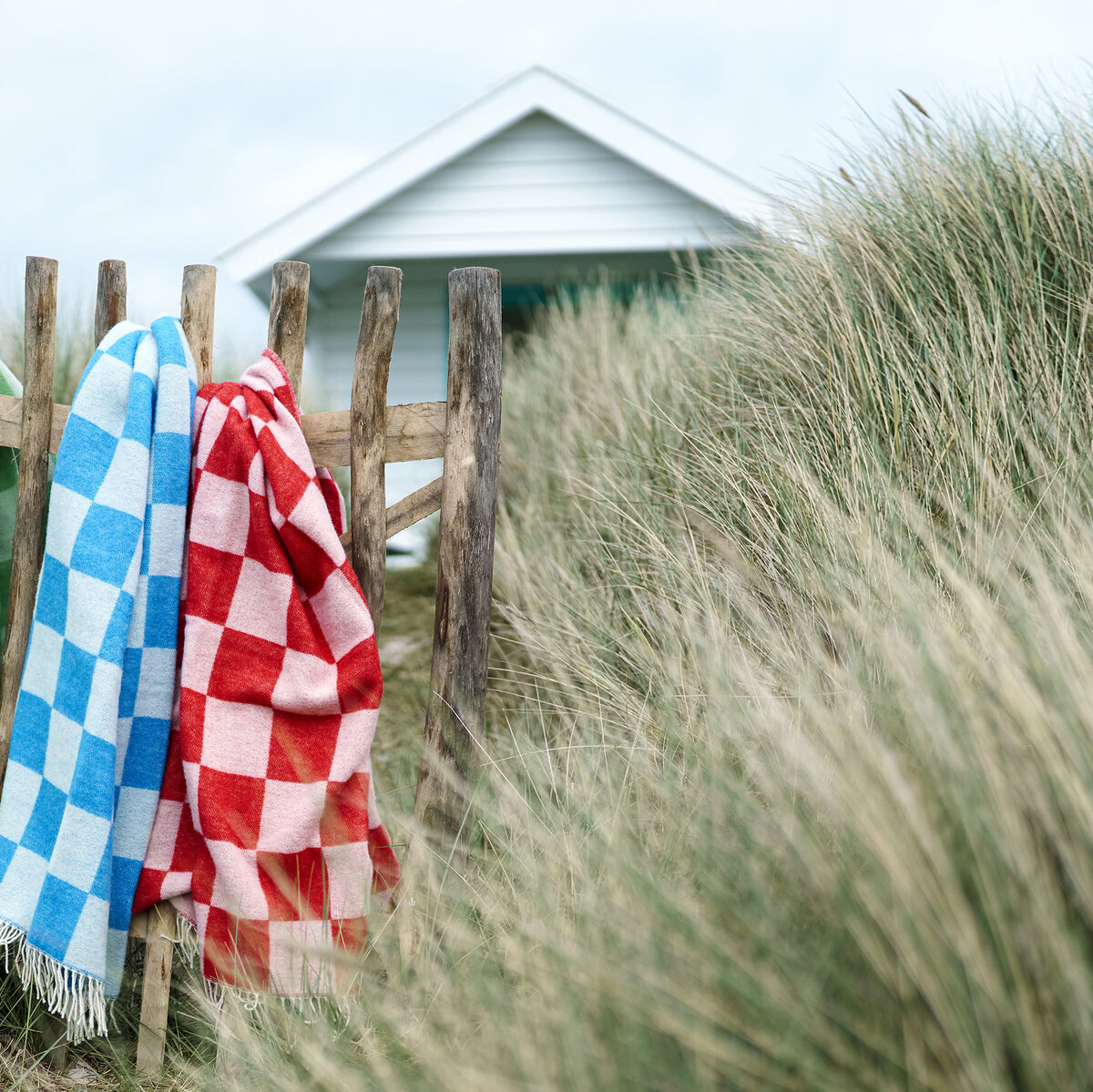 Drei Decken im Karomuster in den Farben Grün, Blau und Rot hängen an einem Holzzaun inmitten von hohen Gräsern nahe einem Strandhaus.