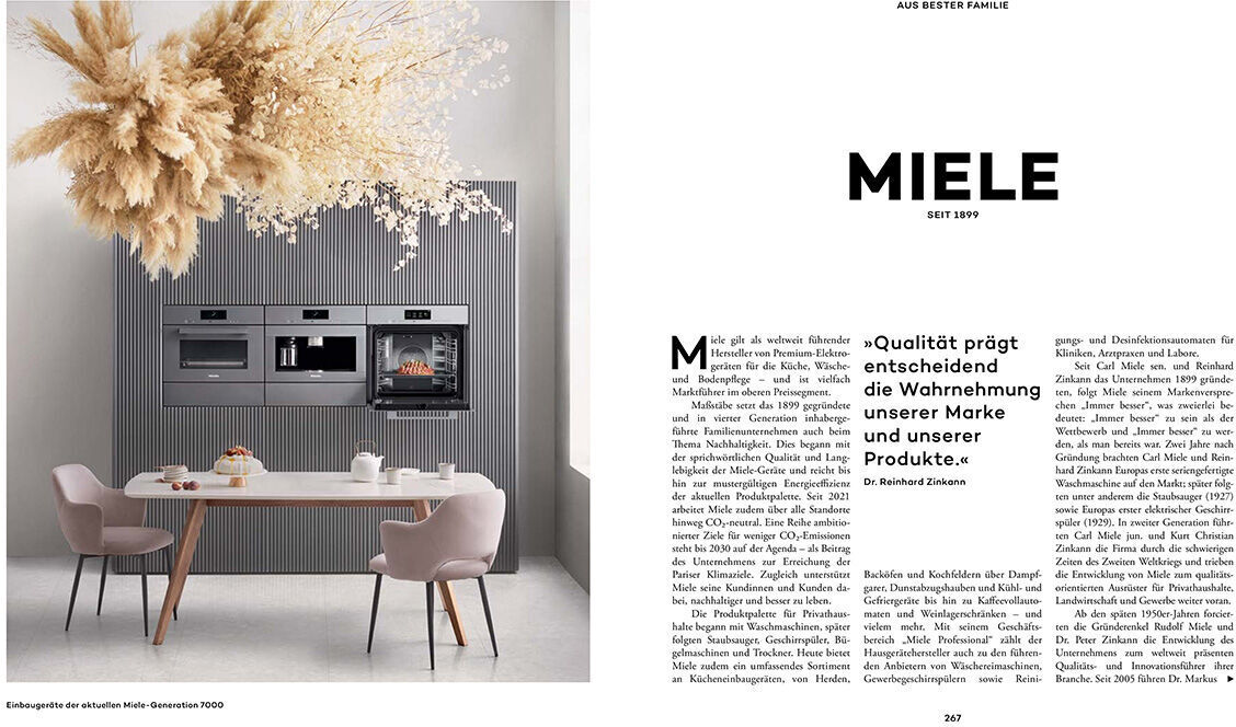 Ein Buch mit dem Titel „Aus bester Familie“ liegt aufgeschlagen auf einem Tisch. Neben dem Buch steht ein moderner Küchenschrank mit eingebauter Miele-Qualität, umgeben von stilvollen Trockenblumen und eleganten, minimalistisch gestalteten Stühlen.
