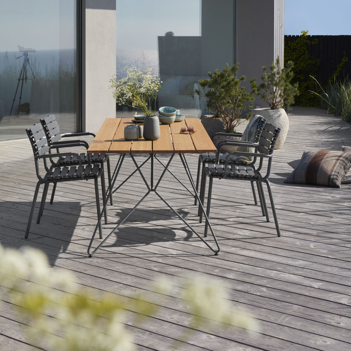 Ein moderner Gartentisch-Set »SKETCH« auf einer Terrasse mit einem Holztisch und vier grauen Metallstühlen. Der Bereich ist geschmackvoll dekoriert mit Pflanzen und Dekorationsstücken auf dem Tisch.