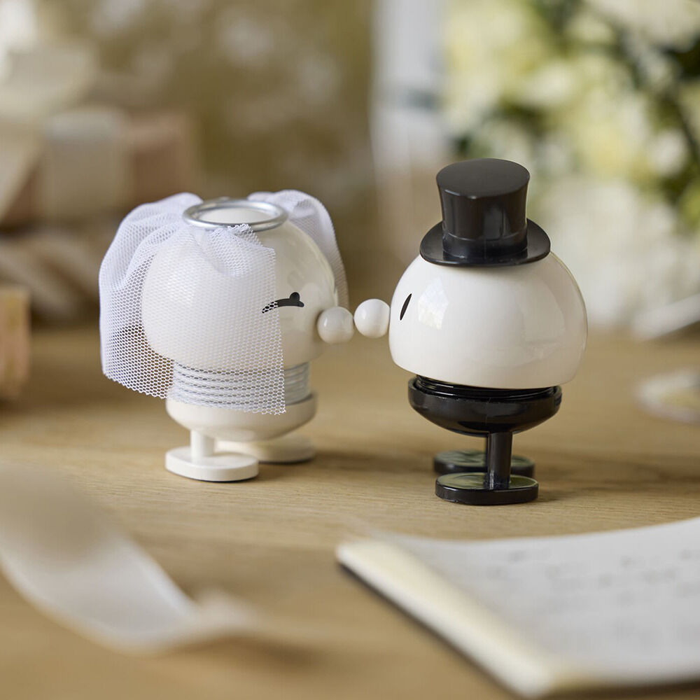 Detailliertes Foto des Hoptimist »Braut & Bräutigam« Sets: Zwei runde, weiße Figuren aus ABS/Stahl, eine im Schleier und eine mit Zylinder, auf einem Holztisch. Im Hintergrund befinden sich Blumen, Geschenke und ein geöffnetes Notizbuch. Moderne, verspielte Dekoration.