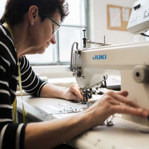 Nahaufnahme einer Person, die an einer Juki-Nähmaschine näht, mit Blick auf ihre Hände, die den Stoff führen. Auf dem Tisch ist ein Maßband zu sehen.