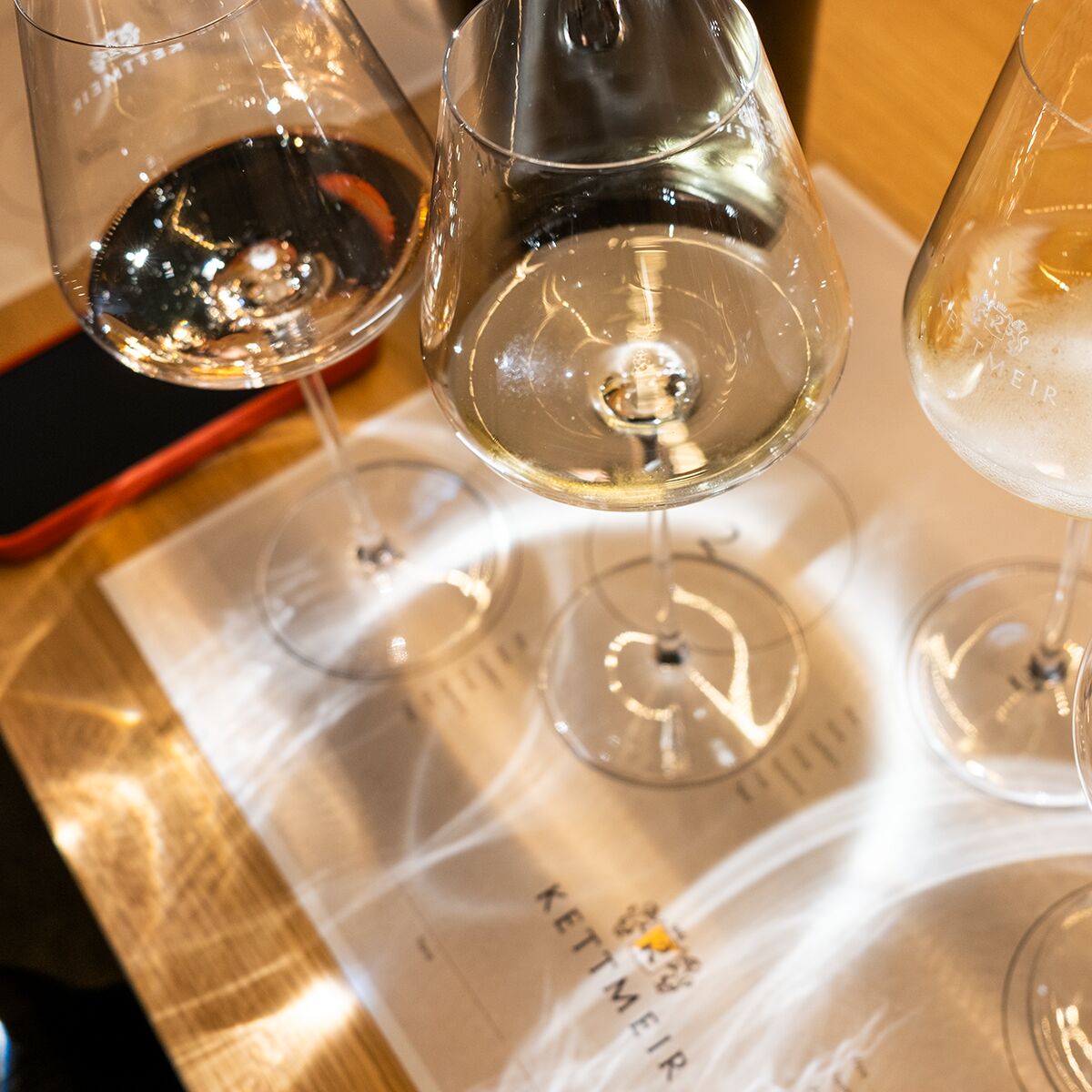 Drei elegante, halbgefüllte Weingläser stehen auf einer hellen Tischdecke mit einem gedruckten Verkostungsbogen der ZEIT-Weinedition »KETTMEIR«. Im Hintergrund schimmern warme Lichtreflexe und teilweise ist ein rotes Smartphone zu sehen.