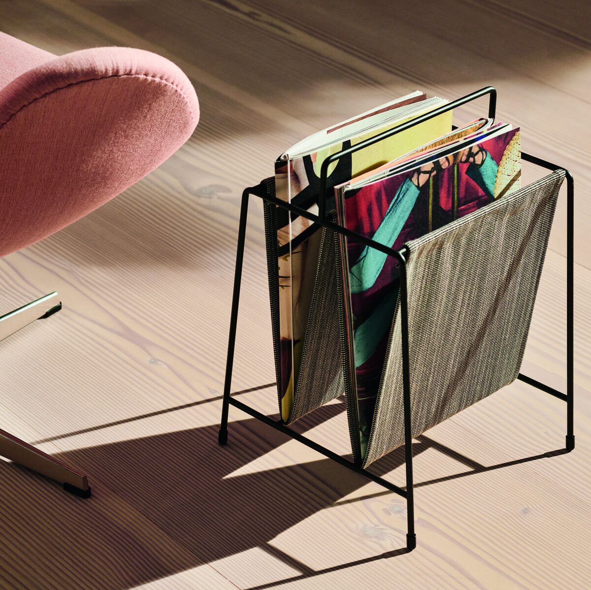 Ein modern gestalteter Zeitungshalter aus schwarzem Stahlrahmen steht auf einem Holzboden neben einem rosa Designerstuhl. Im Halter sind verschiedene Magazine und Zeitschriften abgelegt.
