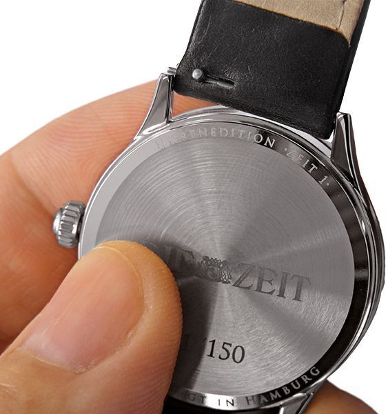 Nahaufnahme einer Armbanduhr aus Edelstahl mit schwarzem Armband und glänzender Rückseite, auf der 'ZEIT 1' eingraviert ist. Eine Hand hält die Uhr.