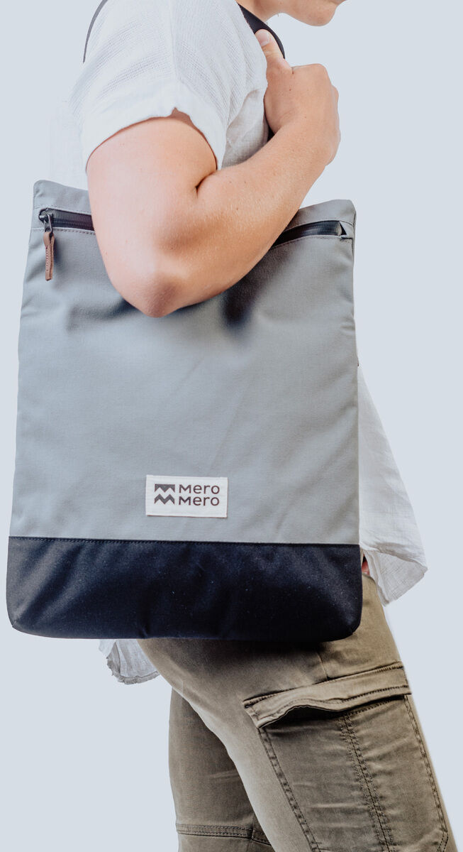 Eine Person trägt eine modische graue Tasche namens »Meije«. Die Tasche hat schwarze und braune Details sowie einen Reißverschluss. Sie ist schlicht, praktisch und stilvoll für den Alltag oder Reisen.