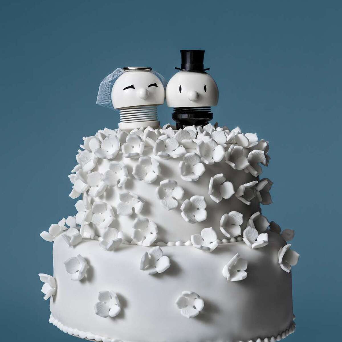 Weiße zweistöckige Hochzeitstorte mit detaillierten weißen Blüten und Braut & Bräutigam Hoptimist-Figuren als Tortenaufsatz. Die Braut trägt Schleier, der Bräutigam Zylinder. Hintergrund in mattem Blau, Gesamtbild modern und stilvoll.