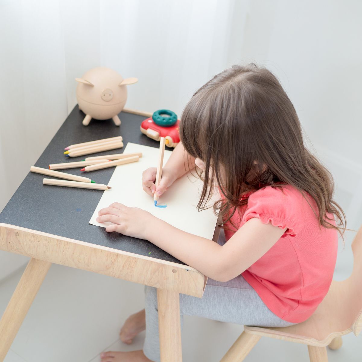 Ein Kind sitzt an einem schwarz-weißen Tisch und malt mit Buntstiften auf einem Blatt Papier. Der Tisch hat Holzbeine und der Stuhl ist ebenfalls aus Holz. Im Hintergrund sind weitere Spielzeuge zu sehen.