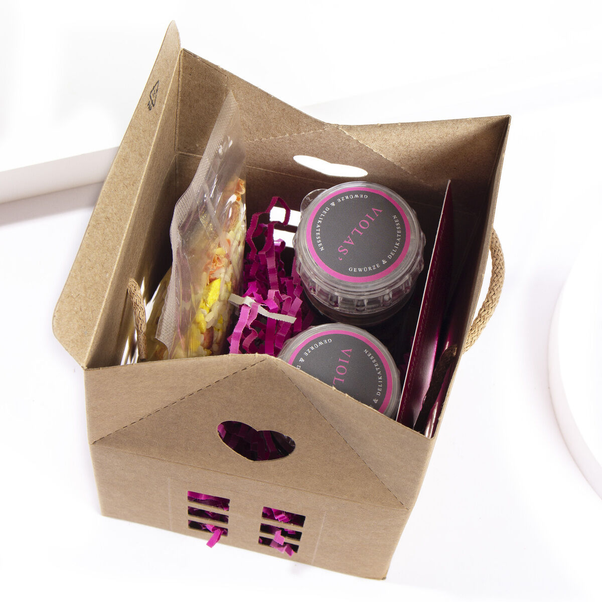 Geschenkset in Form eines kleinen Hauses, enthält Gläser mit Leckereien, dekorative Papiereinschläge und Verpackungsfüllung. Die Farben sind vorwiegend Braun, Rosa und Gelb.