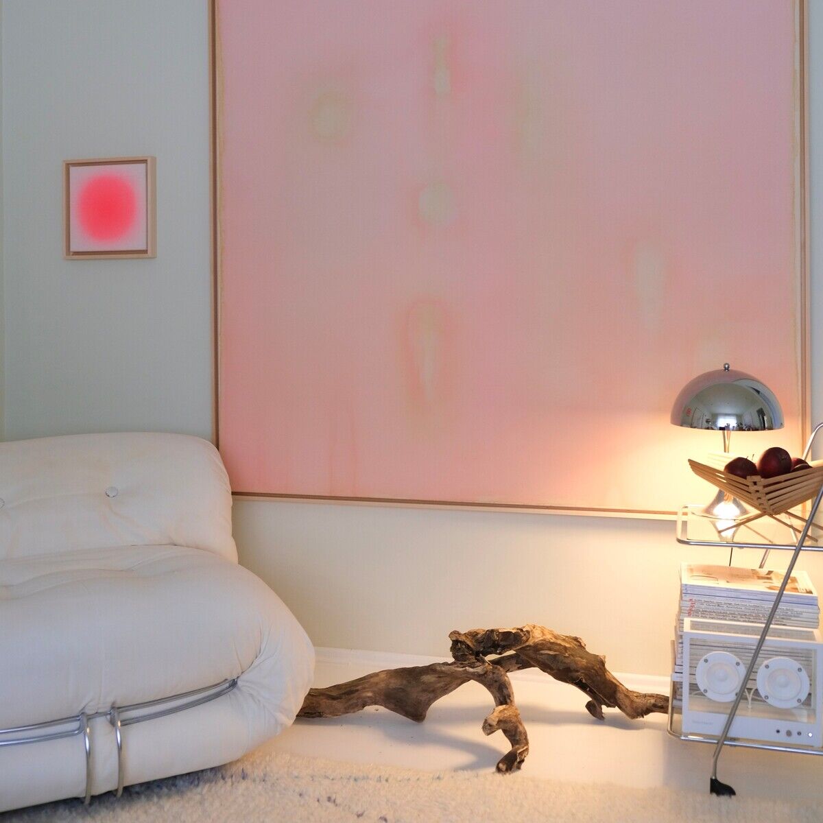 Großes quadratisches Kunstwerk mit roséfarbenen, pastelligen Farbtönen in Holzrahmung hängt an einer hellen Wand über einem modernen weißen Designersofa. Daneben steht eine kleine Lampe auf einem hellen Beistelltisch neben einem Stück gewundenem Treibholz. Die harmonische Szene spiegelt zeitgenössisches und minimalistisches Wohnambiente wider.