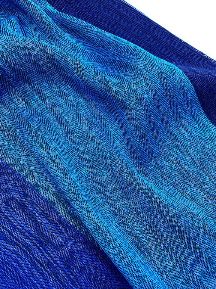 Ein breiter Schal aus blauem Leinen mit einem sanften Farbverlauf von dunkelblau zu hellblau und einem zarten Fischgrätenmuster.