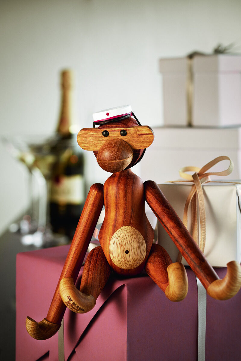 Die Holzfigur »Affe« von Kay Bojesen sitzt auf einem Geschenkstapel. Im Hintergrund sind weitere Geschenke, eine Champagnerflasche und Gläser zu sehen. Die Figur ist aus verschiedenfarbigem Holz gefertigt und trägt ein kleines Mützchen.