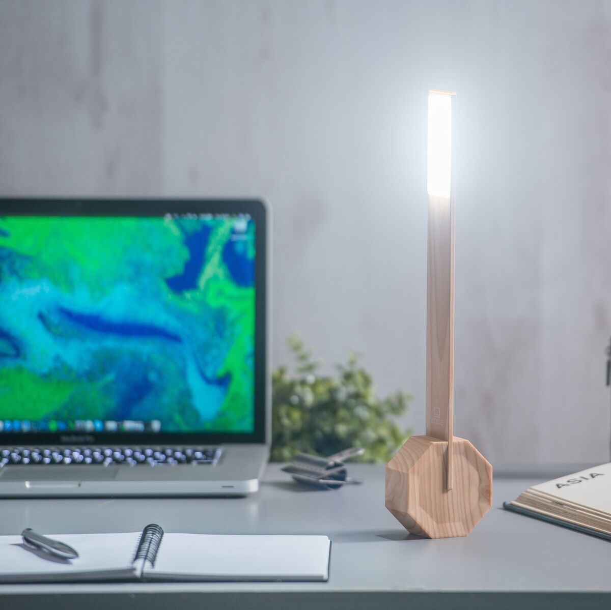 Eine moderne Tischlampe aus Holz, genannt 'Octagon Tischlampe »One«', steht auf einem Schreibtisch. Die Lampe hat ein schlankes, geometrisches Design und leuchtet hell. Im Hintergrund befinden sich ein Laptop, ein Notizbuch und Pflanzen.