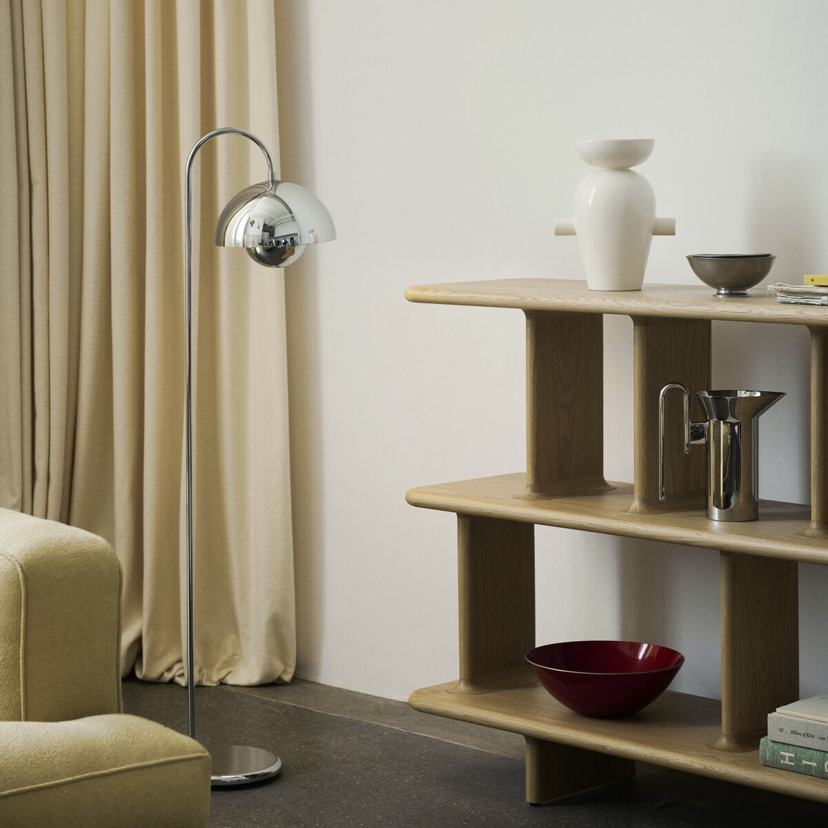 Eine moderne Inneneinrichtungs-Szene mit einem beigen Sofa, einer silbernen Flowerpot VP12 Tischlampe, einem cremefarbenen Vorhang und einem mehrstöckigen Holzregal, das verschiedene Dekorationsgegenstände wie Vasen und Schalen in Cremeweiß und Rot enthält.