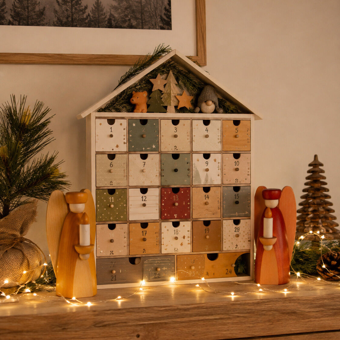 Stimmungsvolles Bild mit beleuchtetem Holz-Adventskalender in Hausform auf Holztisch, dekoriert mit zwei handgefertigten, farbigen Holzfiguren „Lichterengel“ von Björn Köhler aus Lindenholz. Pflanzen, Zapfen und Lichterkette sorgen für festliche Atmosphäre.