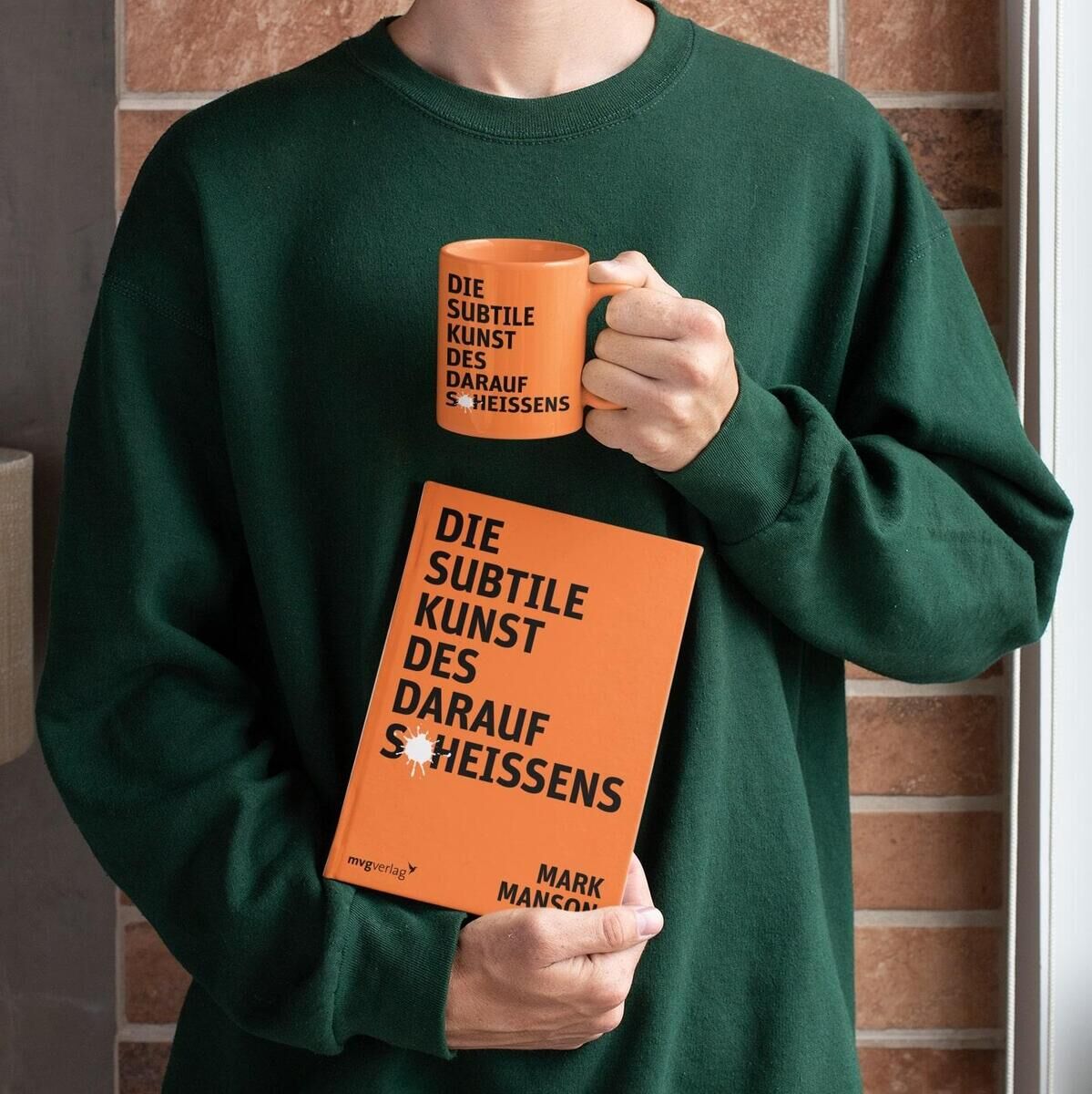 Eine Person hält das Buch 'Die subtile Kunst des darauf Scheißens' von Mark Manson in einer Hand und eine passende orangefarbene Tasse in der anderen Hand. Die Person trägt einen grünen Pullover. Im Hintergrund ist eine beigen Kachelwand zu sehen.
