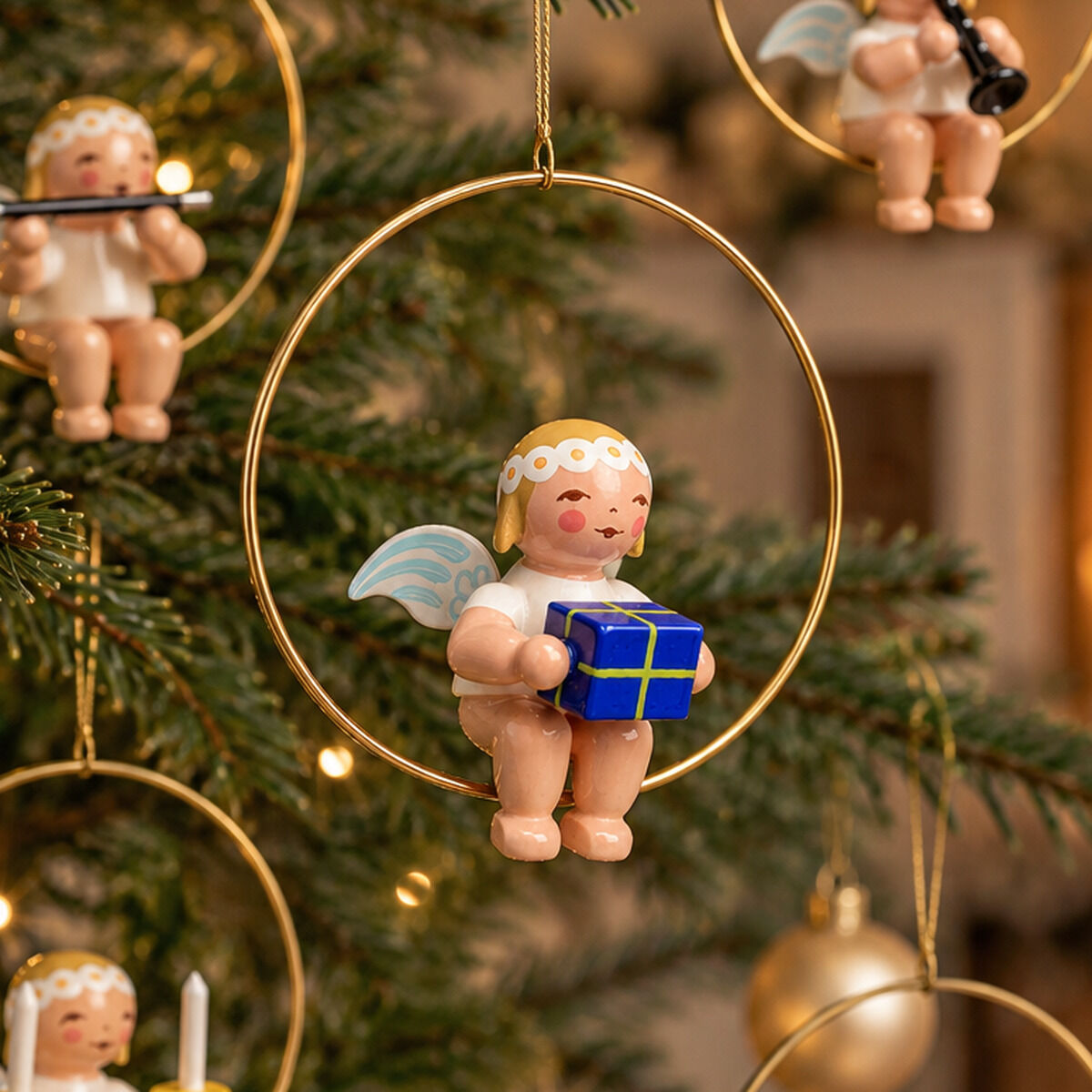 Fünf handbemalte Holzfiguren von Wendt & Kühn, Christbaumengel im Ring, hängen an einem festlich geschmückten Weihnachtsbaum. Jeder Engel hält ein Geschenk oder ein Instrument. Die Szenen sind von warmem Licht und goldenem Baumschmuck umgeben.