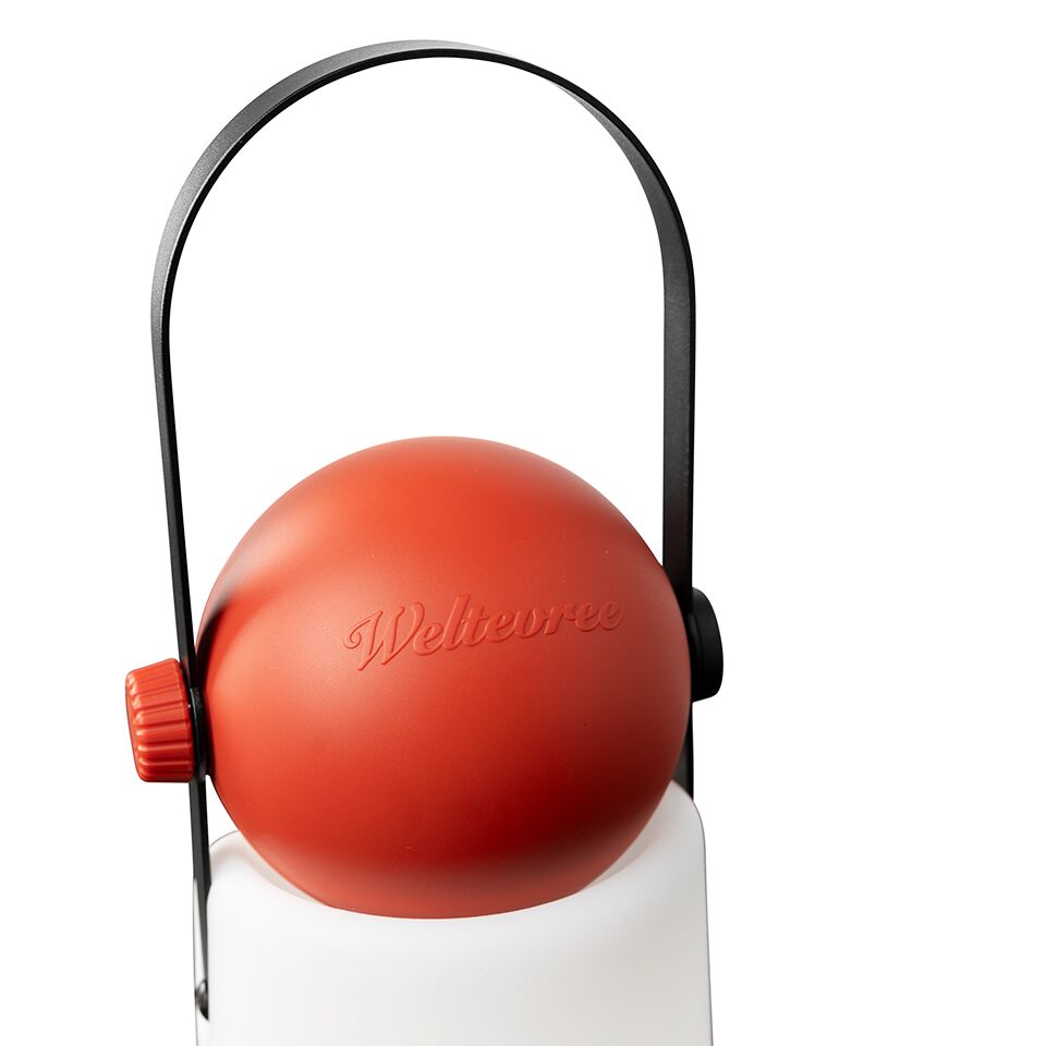 Die Abbildung zeigt die tragbare Leuchte "Guidelight" der Marke Weltevree. Sie besitzt einen weißen zylindrischen Lampenkörper, einen auffälligen roten Drehknopf und eine rote, kugelförmige Abdeckung mit eingeprägtem Markenschriftzug. Ein schwarzer Bügel dient als Griff oder Aufhängung.