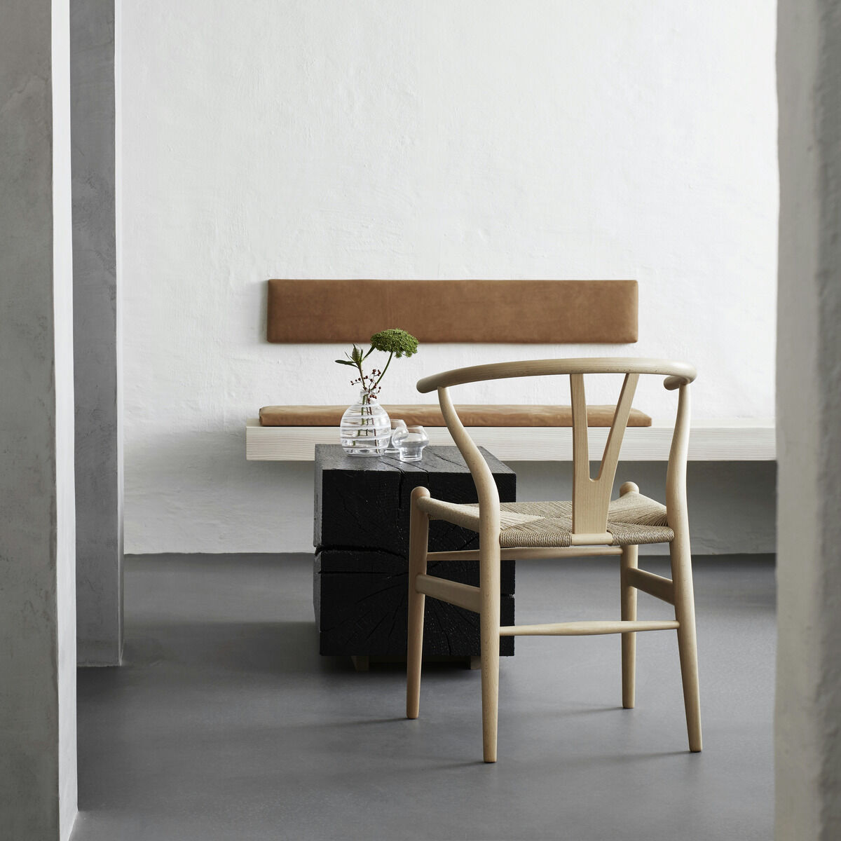 Im Bild steht ein Wishbone Chair »CH24« von Carl Hansen & Søn aus Buche geseift im Fokus eines modernen, minimalistisch eingerichteten Raumes mit weißen Wänden, grauem Boden und schlichter Sitzbank. Auf dem Tisch stehen Glasvasen mit einer Blume.