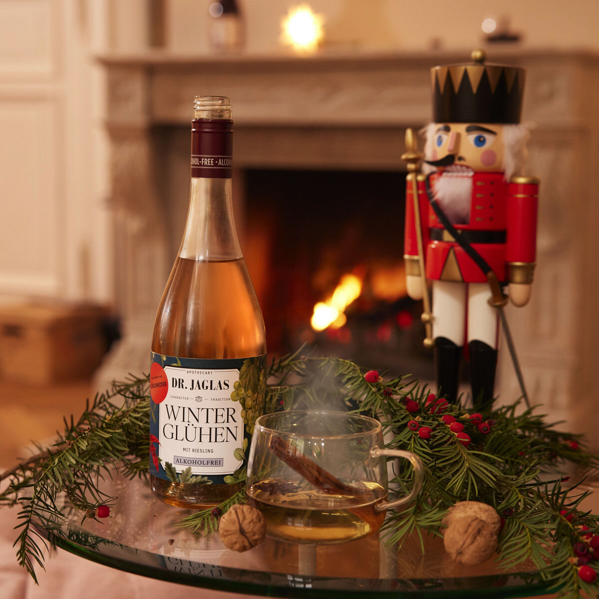 Eine Glasflasche DR. JAGLAS Winterglühen Riesling alkoholfrei steht auf einem Glastisch, umgeben von Tannenzweigen, roten Beeren, Nüssen und einem Glas mit Zimtstange. Im Hintergrund brennt ein Kamin, daneben steht ein großer Nussknacker als Dekoration.