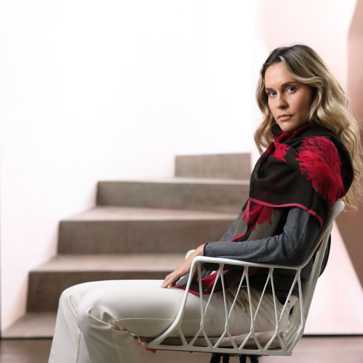 Eine junge Frau sitzt auf einem modernen Stuhl und trägt einen roten Wollschal namens 'Carol', kombiniert mit einem grauen Oberteil, weißen Hosen und schwarzen Sneakers. Der Hintergrund ist minimalistisch mit einer Steintreppe, das Ambiente wirkt modern und stilvoll.