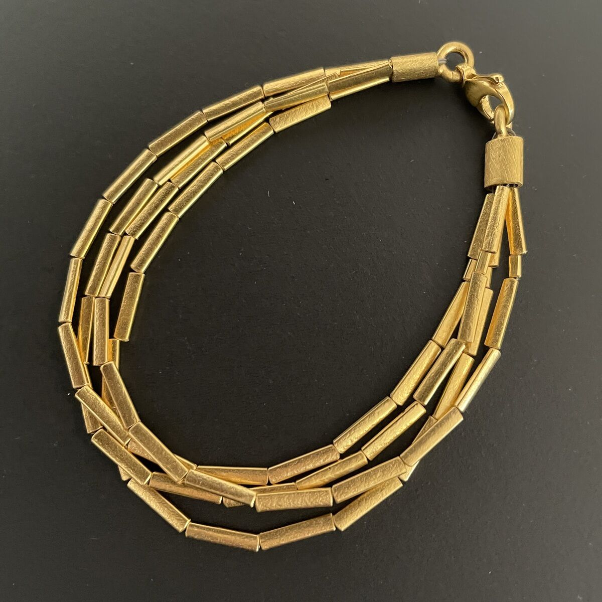 Dreireihiges Armband mit feinen, zylindrischen Gliedern aus Silber, feingoldplattiert, auf schwarzem Hintergrund, modernes und elegantes Schmuckstück für besondere Anlässe.