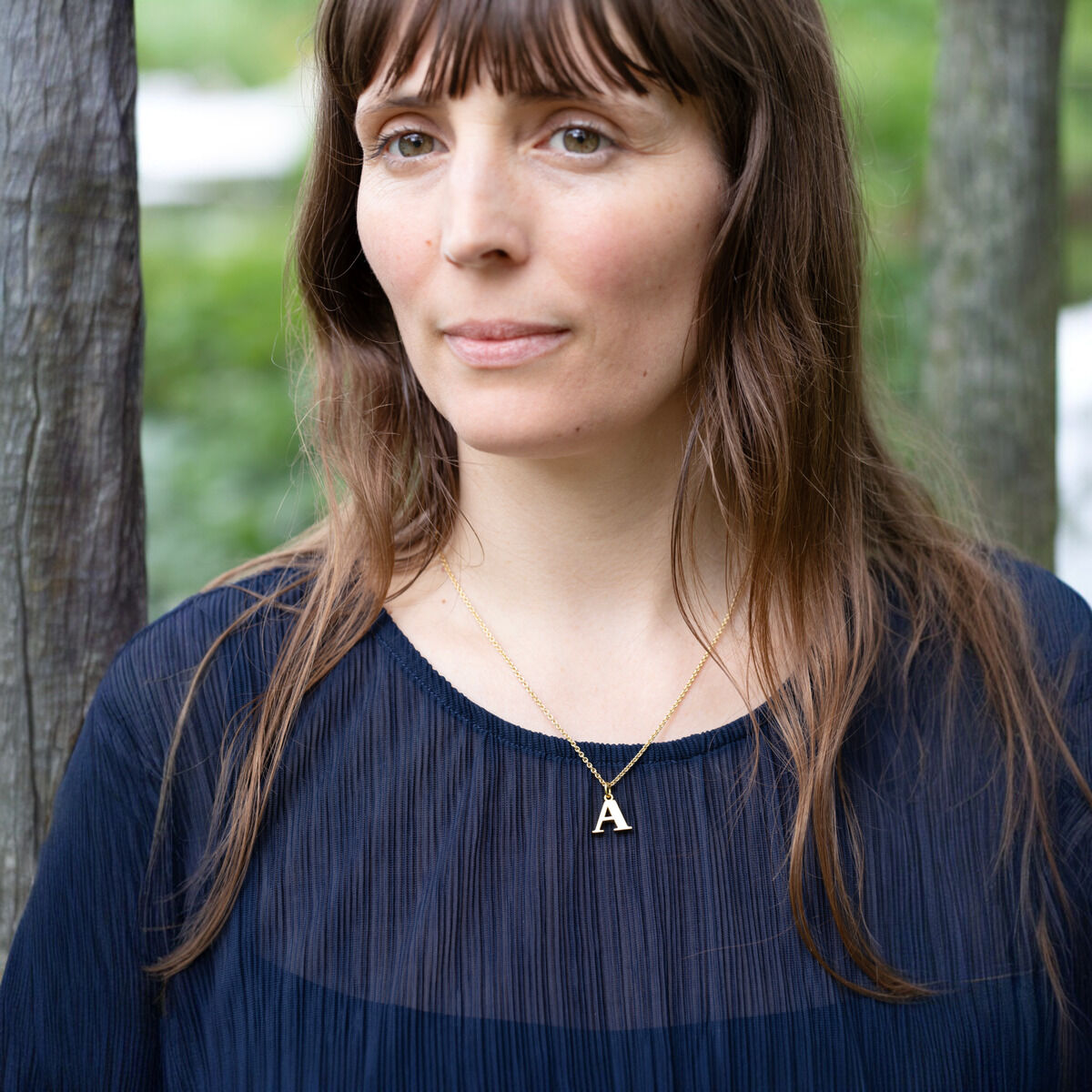 Das Bild zeigt eine Frau, die einen vergoldeten Buchstabenanhänger in Form des Buchstabens 'A' an einer Kette trägt. Sie hat langes braunes Haar und trägt ein dunkelblaues Oberteil, während sie im Freien steht.