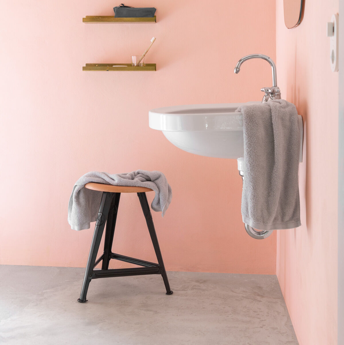 Modern gestaltetes Badezimmer mit einem durchdachten minimalistischen Design. Der Rowac-Schemel in der Holzart Eiche mit schwarzem Gestell steht unter einem mit einem Handtuch bedeckten Waschbecken. An der Wand hängen minimalistische Regale mit einigen dezenten Dekoelementen. Die Wände sind in einem warmen Rosa gehalten.