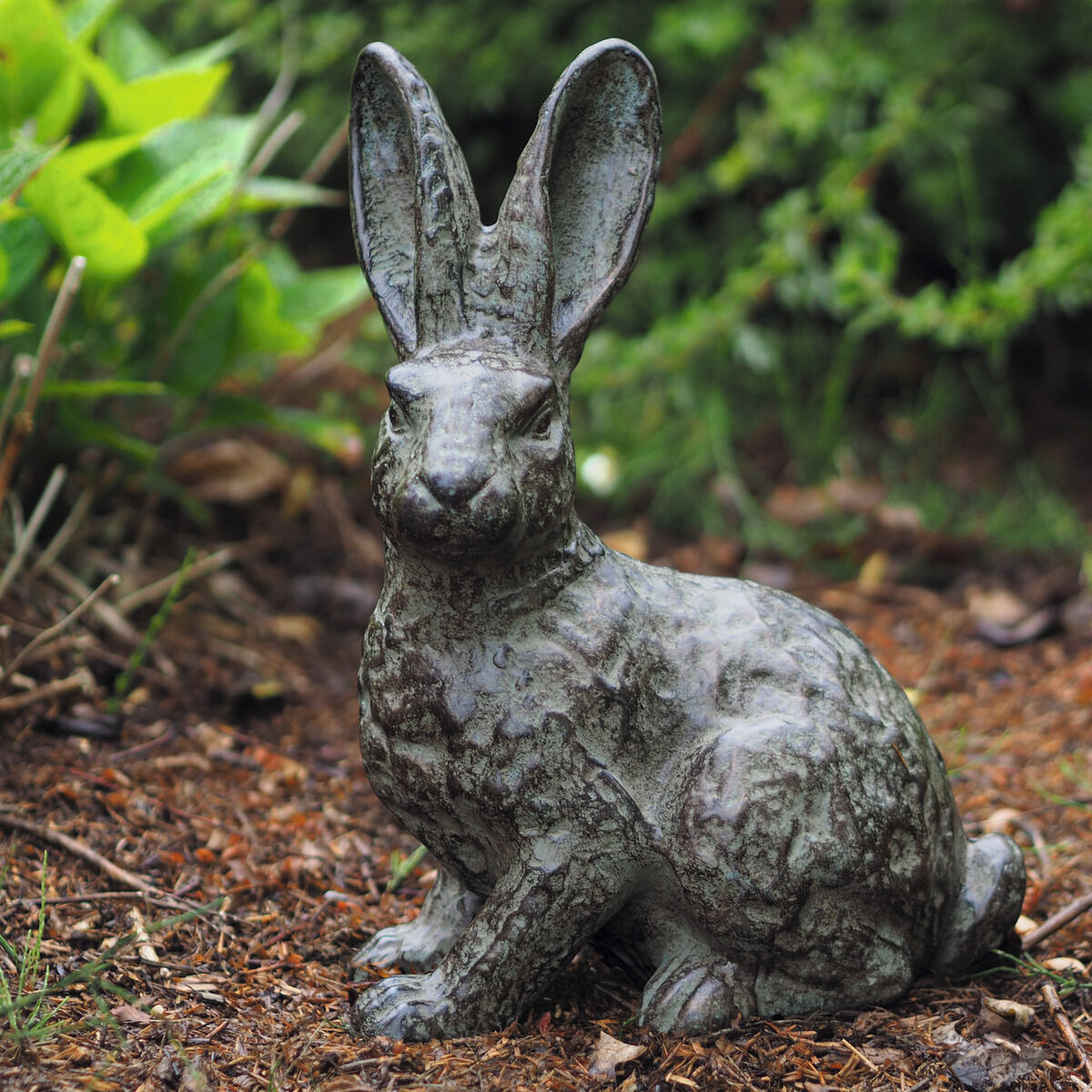Bronzefarbene Gartenskulptur eines sitzenden Hasen mit aufgerichteten Ohren auf Waldboden, umgeben von natürlichem grünen Bewuchs. Die Oberfläche zeigt feine Patina, das Material wirkt schwer und detailreich gearbeitet. Sehr natürliche Darstellung.