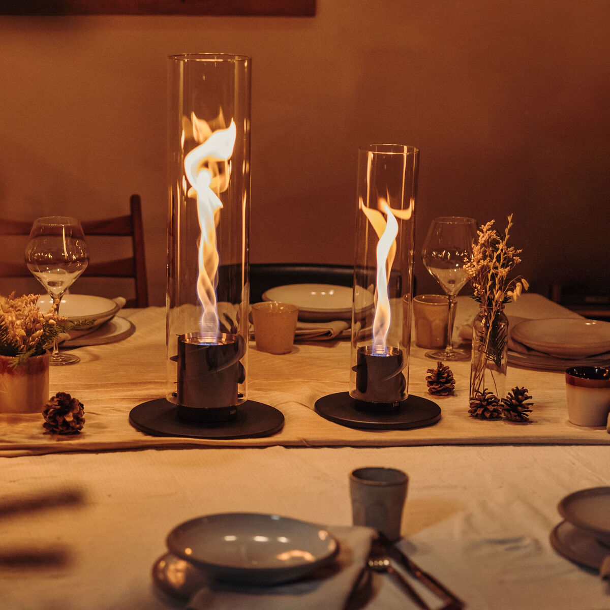 Gemütlich gedeckter Tisch mit Geschirr und zwei brennenden Tischfeuern (SPIN 900) unter Glaszylindern im Innenraum. Die warmen Lichtstrahlen der Flammen sorgen für eine stimmungsvolle Beleuchtung.