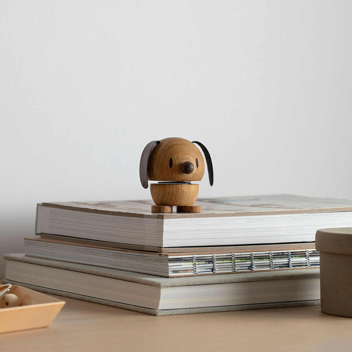 Eine Hoptimist-Figur aus Eichenholz in Form eines Hundes, platziert auf einem Stapel Bücher. Die Figur hat ein minimalistisches und humorvolles Design, ideal als Dekoration oder Geschenk.