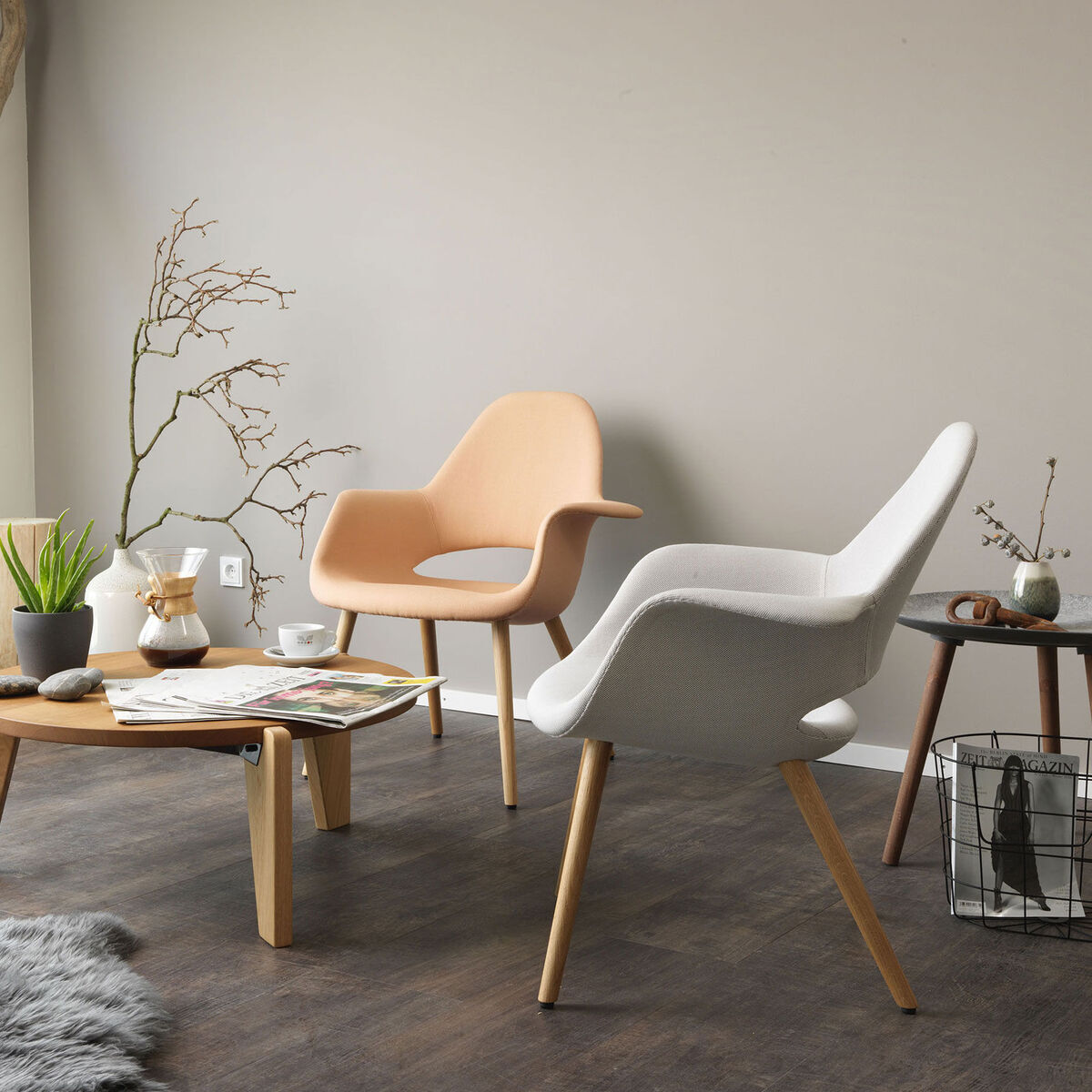 Modernes Wohnzimmer mit zwei ZEIT-Sonderedition »Organic Chair« von Vitra. Ein beiger und ein weißer Stuhl mit Holzbeinen, umgeben von minimalistischen und natürlichen Dekorationselementen. Ein runder Holztisch mit einer Pflanze und Zeitschriften steht in der Mitte des Raumes neben einem Fenster.