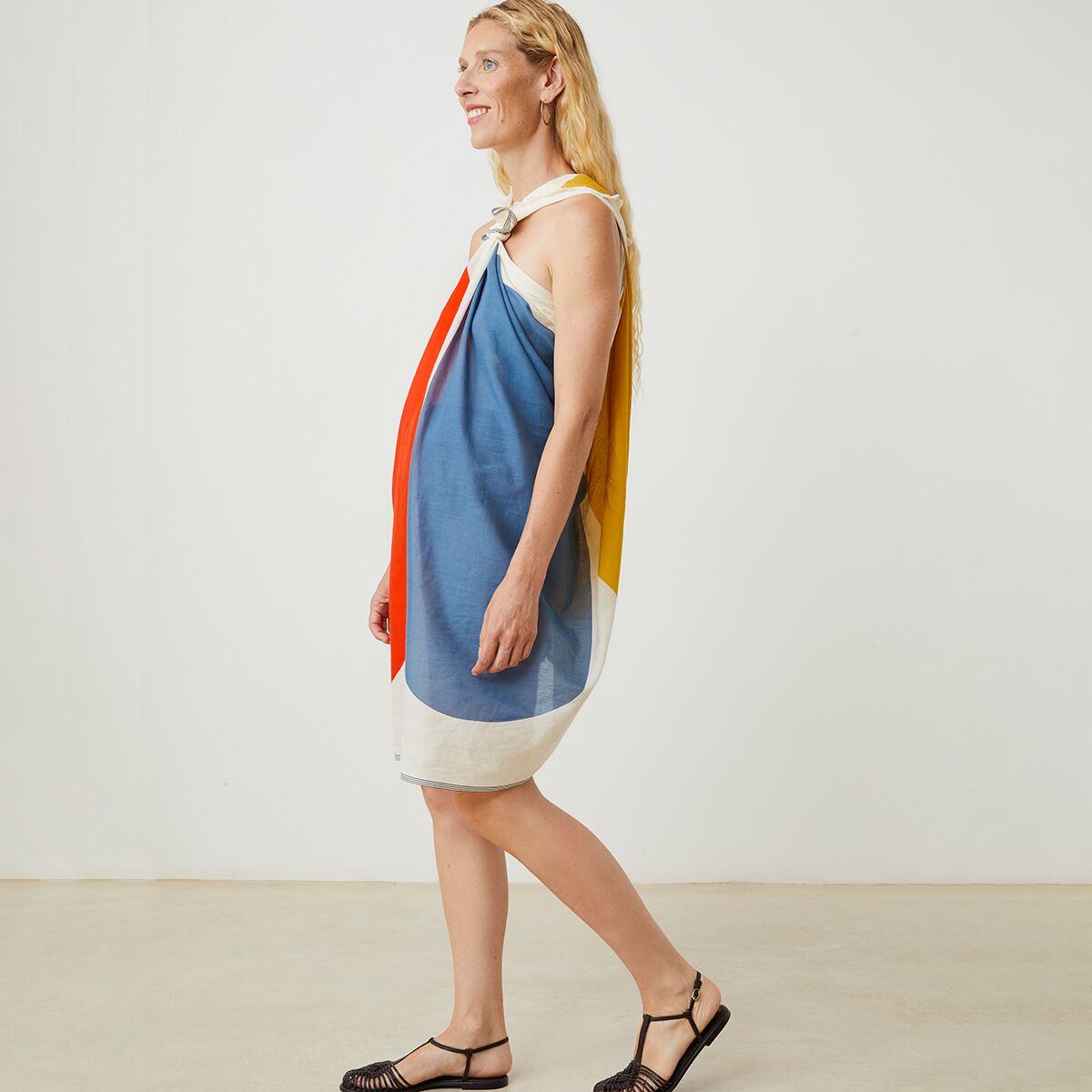 Eine Frau trägt den Baumwollschal »No. 727« von MOISMONT locker um die Schultern gebunden als Kleid. Der Schal zeigt große Farbflächen in Blau, Weiß, Rot und Gelb. Der Hintergrund ist neutral, der Fokus liegt auf dem farbenfrohen, leichten Schal.