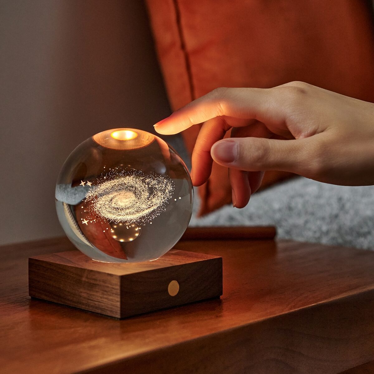 Eine dekorative Amber Light Lampe mit einer gläsernen Kugel auf einem Holzsockel, die ein leuchtendes Galaxie-Muster zeigt. Die Lampe steht auf einem Holztisch in einem gemütlich eingerichteten Raum, und eine Hand schaltet die Lampe ein oder aus.