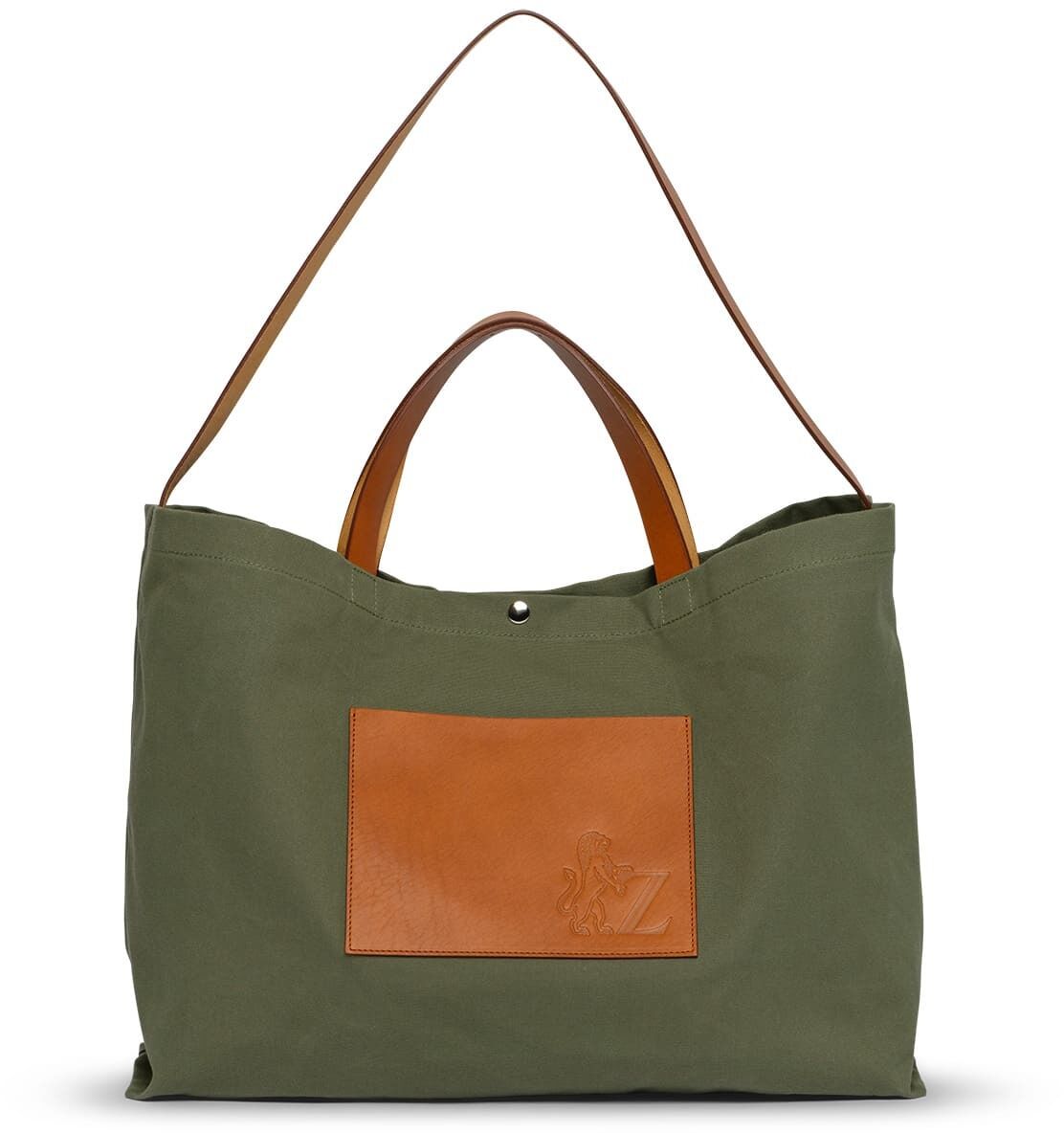 Eine grüne Tragetasche mit braunen Lederdetails und einem Außenfach aus Leder. Die Tasche hat sowohl kurze Henkel als auch einen langen Schulterriemen.