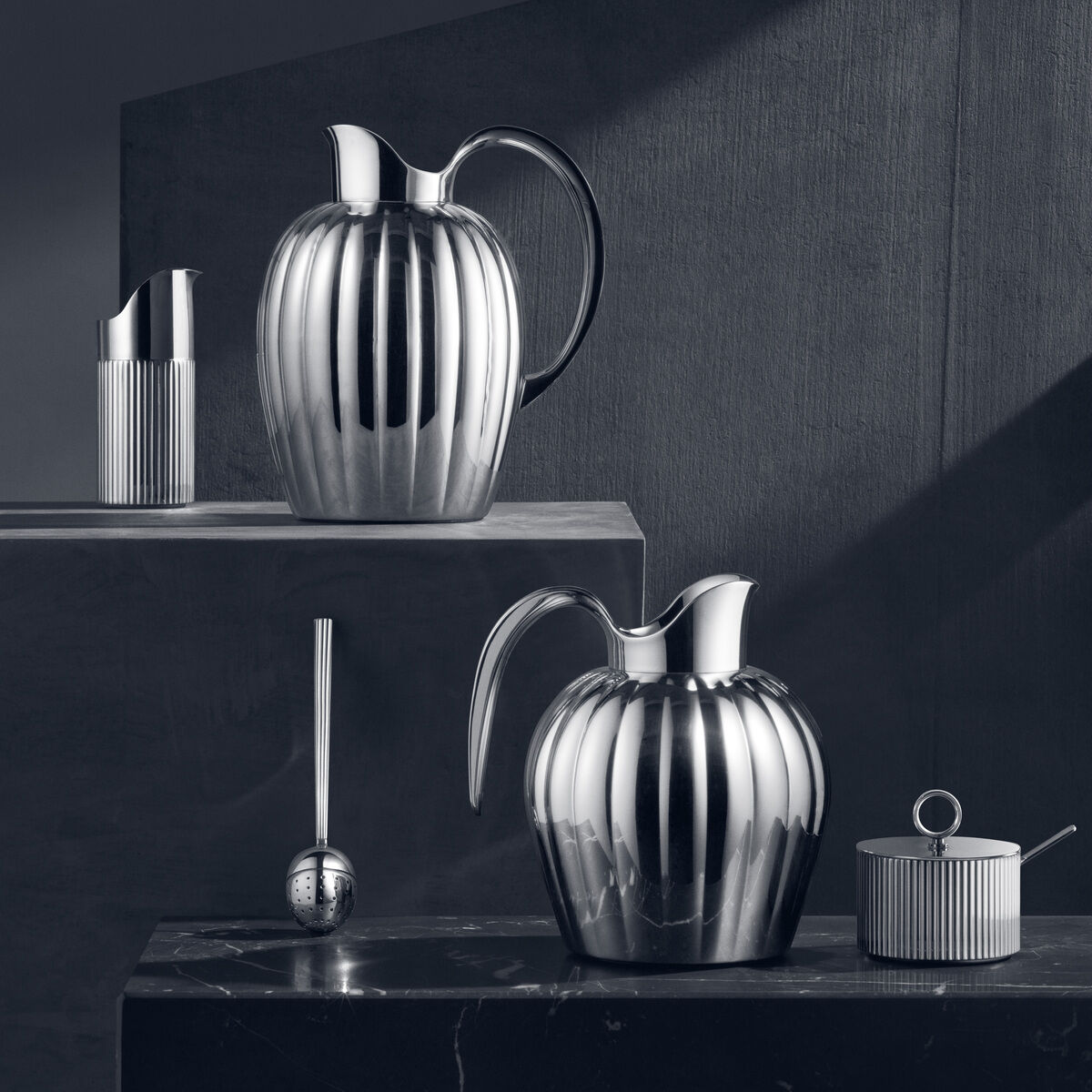 Ein stilvolles Sahnekännchen und passendes Teeservice der Serie »Bernadotte« aus glänzendem Edelstahl, fotografiert in einer ästhetischen, modernen Umgebung, betont durch einen Hintergrund in dunklen Grautönen.