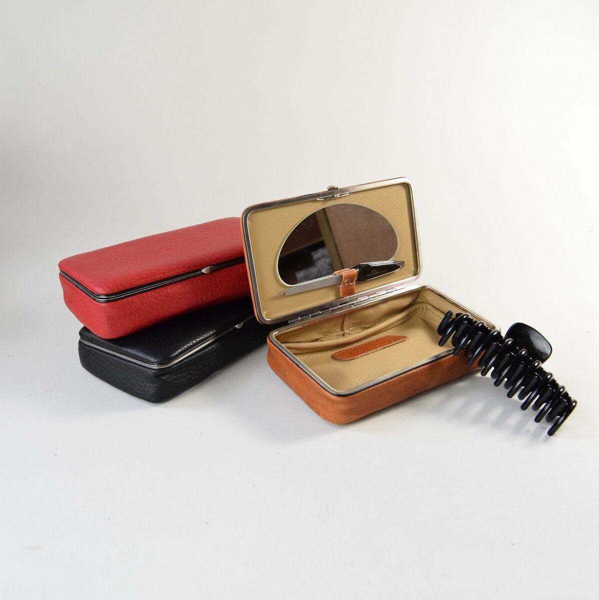 Eine Utensilienbox aus Leder in verschiedenen Farben, darunter Schwarz, Rot und Braun. Die Boxen haben jeweils einen Reißverschluss, und eine geöffnete Box zeigt ein beige Innenfutter mit Haarspangen und einem Spiegel.