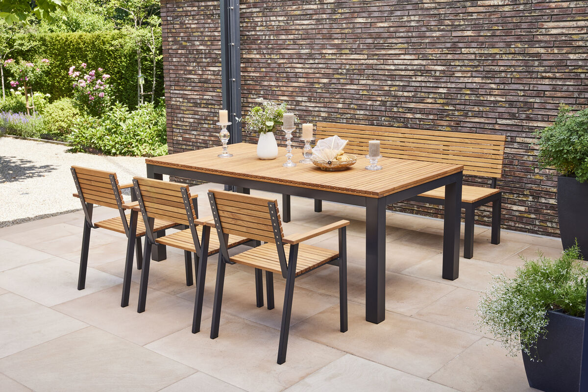 Gartenmöbelsatz mit einem Tisch, vier Stühlen und einer 2er Bank aus Holz und schwarzem Metall auf einer Terrasse, umgeben von grünen Pflanzen.