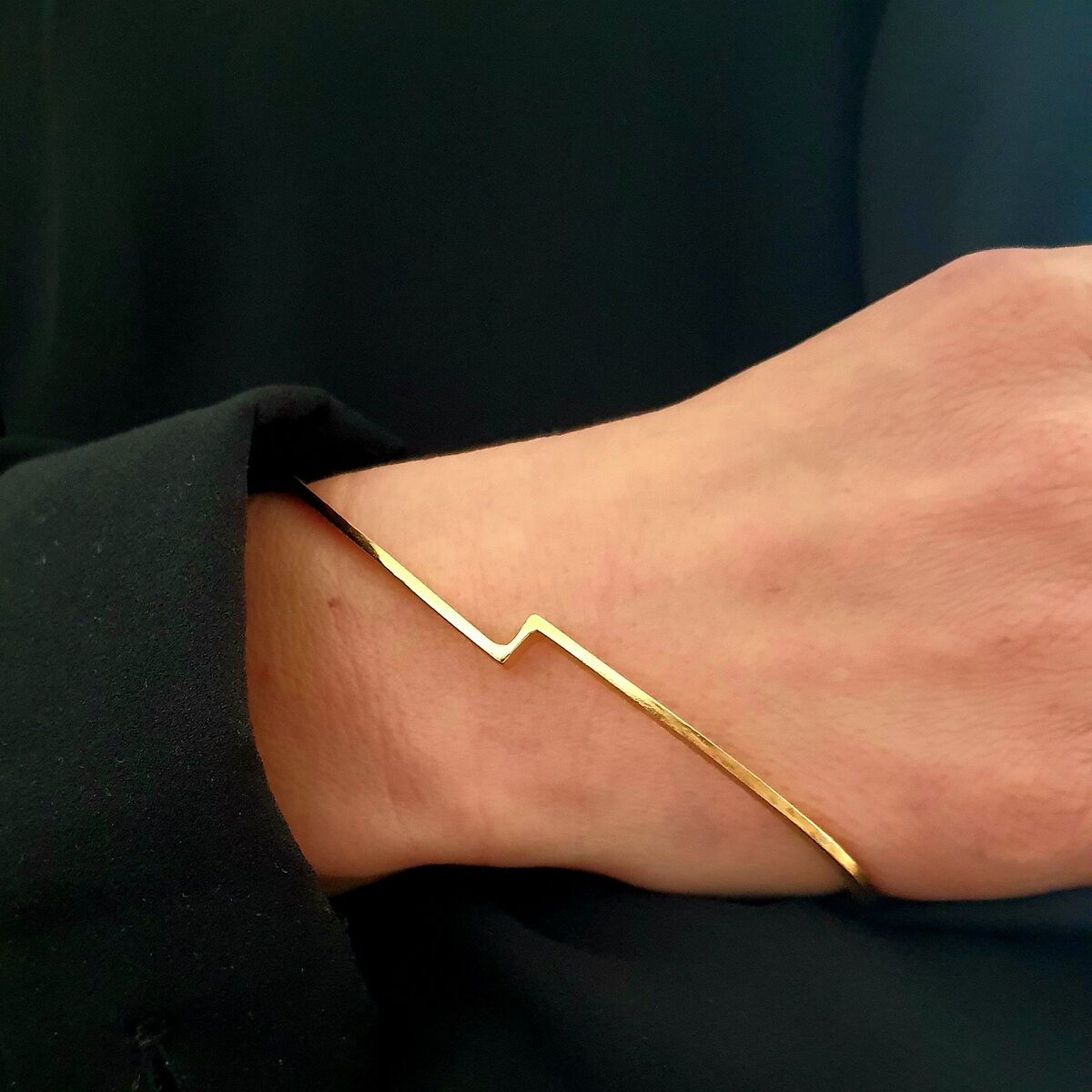 Ein goldener Armreif mit einem minimalistischen Design wird von einer Person getragen, die schwarze Kleidung anhat. Der Armreif ist schlicht und elegant.