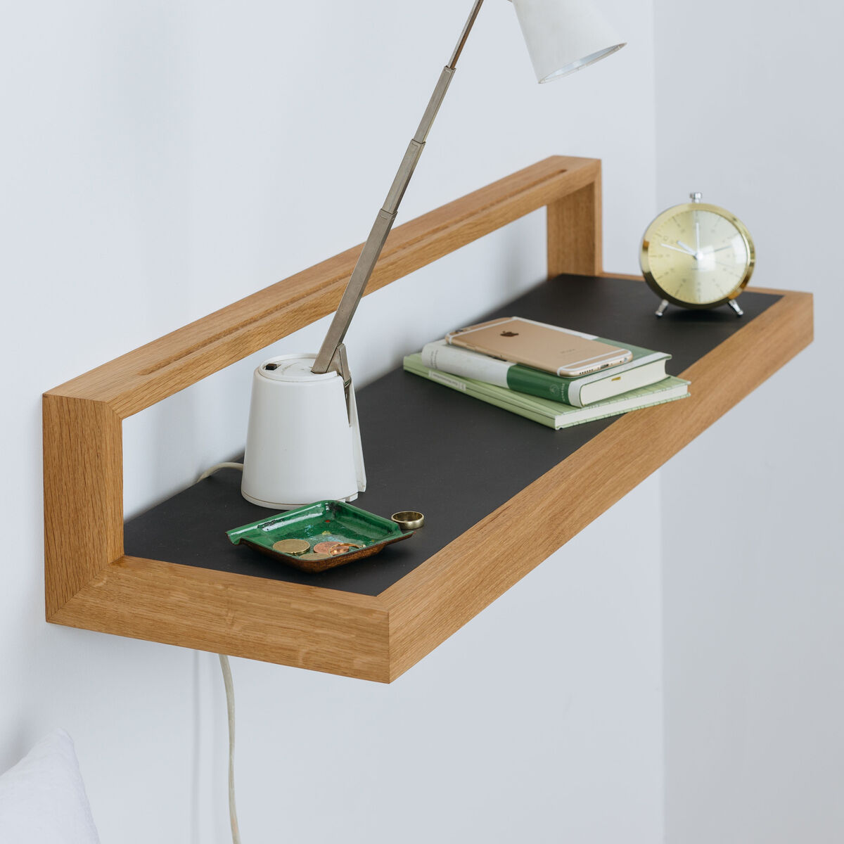 Das Ablagebord »buck« aus geölter Eiche mit anthrazitfarbener Linoleumfläche ist minimalistisch gestaltet und an der Wand montiert. Auf dem Regal stehen eine weiße Schreibtischlampe, ein grünes Tablett, Bücher, ein Notizbuch und ein runder, goldfarbener Wecker.