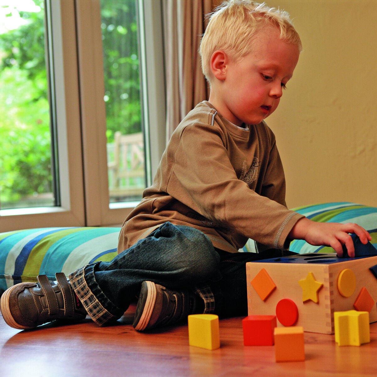 Ein kleines Kind sitzt auf dem Boden und spielt mit einer Sortierbox. Die Sortierbox und die herumliegenden Formen sind farbenfroh in Blau, Gelb, Rot und Orange. Im Hintergrund sind bunte Kissen und eine verglaste Tür zu sehen.