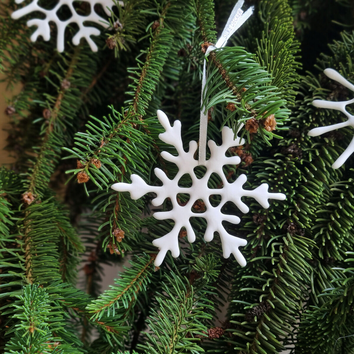 Das Bild zeigt ein Set aus sechs weißen Porzellan-Schneekristallen in verschiedenen Größen, die an grünen Tannenzweigen aufgehängt sind. Die Schneeflocken haben filigrane, handgefertigte Designs.