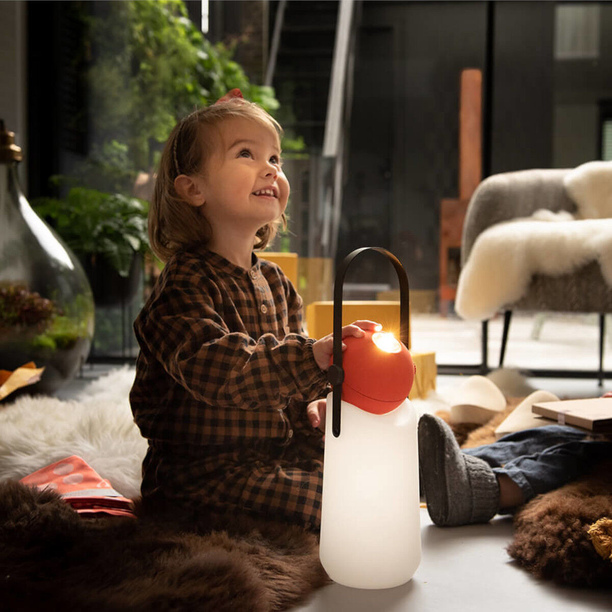 Ein lächelndes Kind im Kinderzimmer hält eine tragbare, weiße LED-Lampe mit einem orangefarbenen Griff. Im Hintergrund sind Pflanzen und Möbel zu sehen.