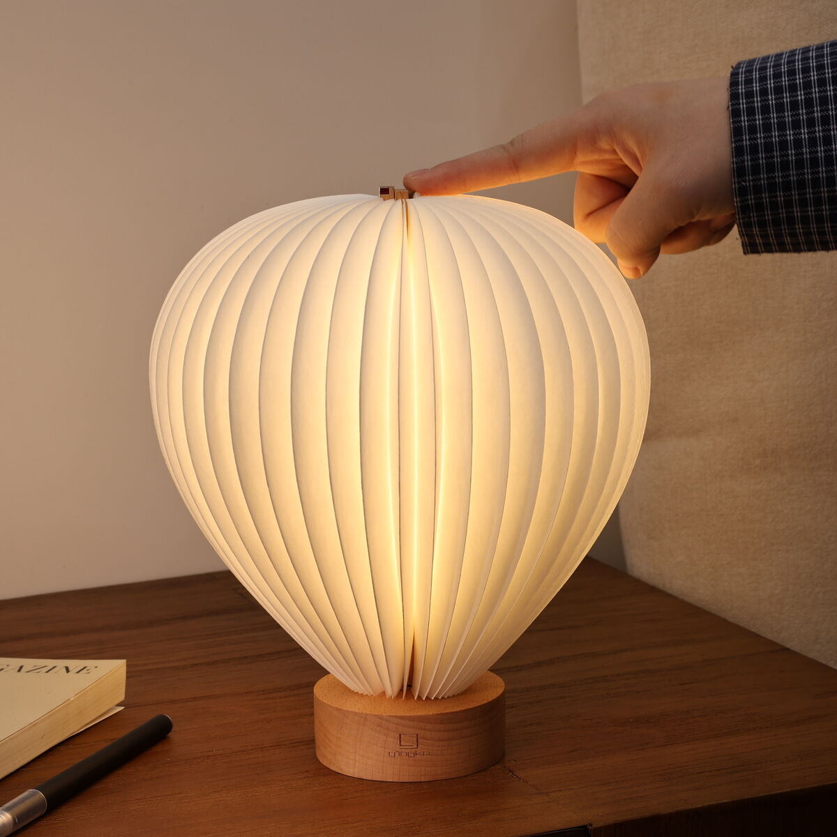 Auf einem Holz-Nachttisch steht die Designerleuchte »Aeris Light« von Gingko Design Ltd in Form eines Heißluftballons mit weißem, gefächertem Lampenschirm aus Papier und einem Fuß aus Holz. Eine Hand schaltet die leuchtende Lampe ein. Daneben liegen ein Buch und ein Stift.