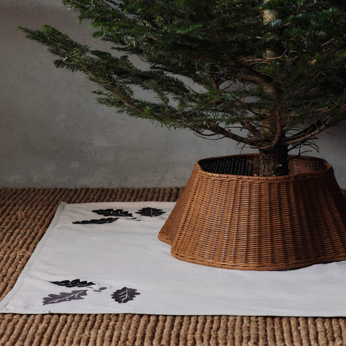 Auf dem Bild ist eine Christbaumdecke mit schwarzen Eichenblättern auf weißem Stoff unter einem Weihnachtsbaum zu sehen. Der Baum steht in einem braunen, geflochtenen Korb auf einem naturfarbenen Teppich in einem schlichten Raum.