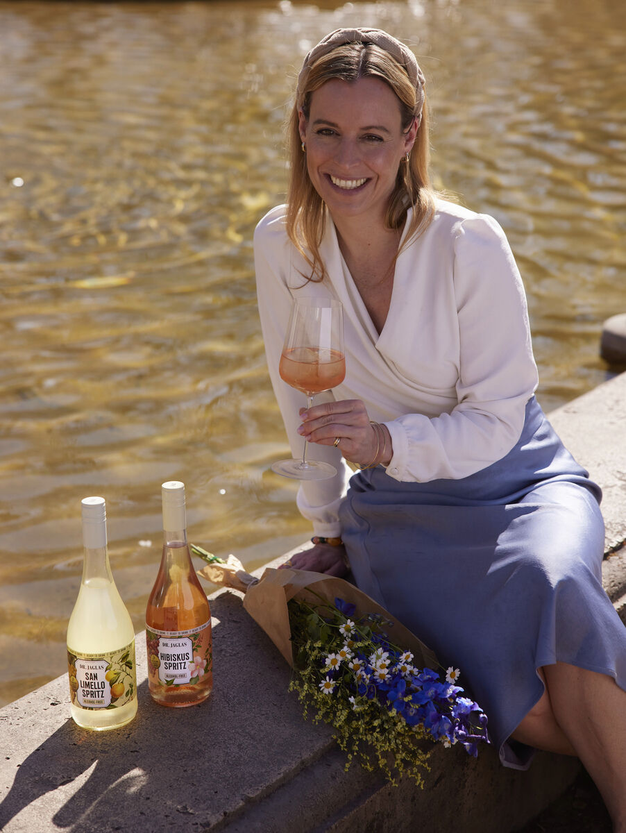 Eine Frau sitzt draußen auf einem Steinsockel vor einem Brunnen und hält ein Glas mit einem orangefarbenen Getränk. Neben ihr stehen zwei Flaschen Hibiskus Spritz und ein Blumenstrauß mit blauen und gelben Blumen.