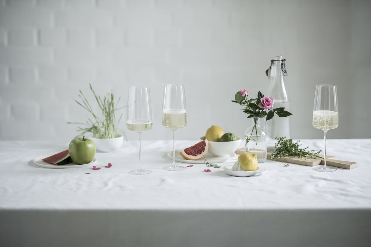Ein Set von zwei eleganten Sektgläsern mit Inhalt steht auf einem Tisch. Der Tisch ist dekoriert mit verschiedenen Früchten, Blumen und Kräutern. Im Hintergrund ist ein helles, minimalistisches Design zu sehen.