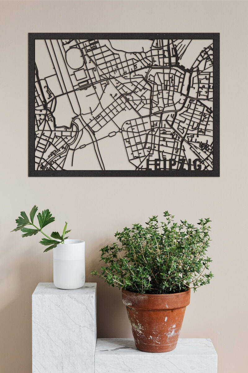 Ein minimalistischer Holzschnitt der Stadtkarte von Leipzig, eingerahmt und an einer beigefarbenen Wand aufgehängt. Darunter stehen auf einem weißen Marmorpodest ein Blumentopf mit Kräuterpflanze und eine weiße Vase.