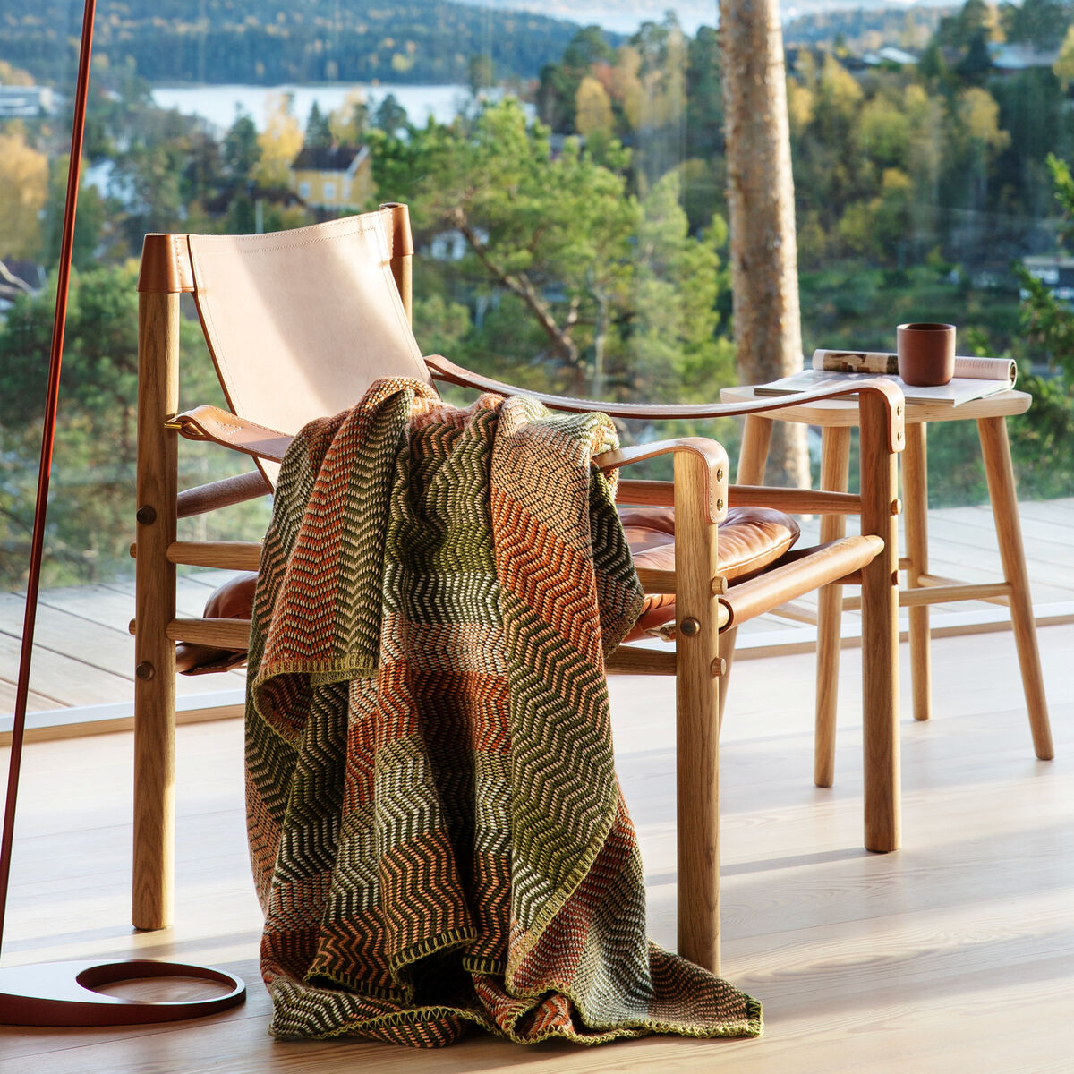 Eine beige-orange-grüne Lammwolldecke liegt auf einem stilvollen Holzstuhl in einem modernen Wohnzimmer mit großen Fenstern. Ein Tisch mit einer Tasse und Büchern steht daneben. Die Natur draußen bietet eine ruhige Kulisse.