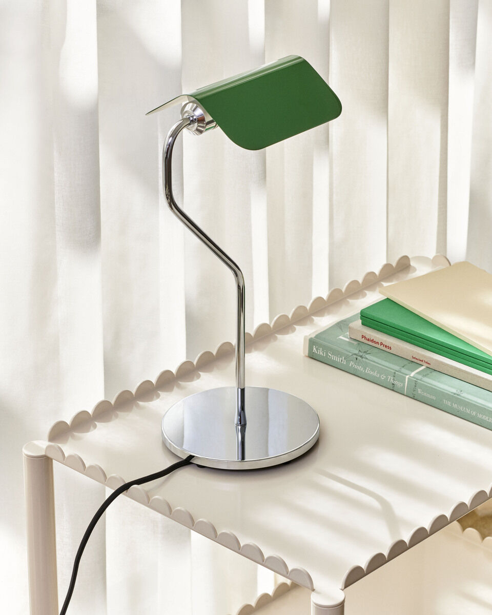Eine moderne Tischlampe mit einem grünen Lampenschirm und einem eleganten gebogenen Edelstahlarm steht auf einem kleinen Tisch. Im Hintergrund sind geschlossene weiße Vorhänge zu sehen.